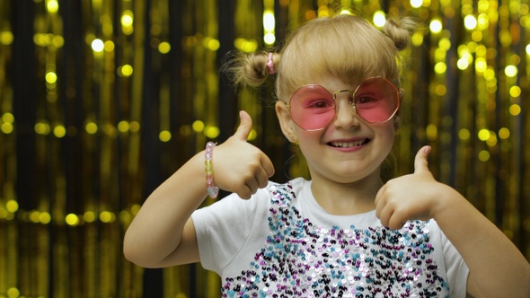 Child Show Thumbs Up, Smiling, Looking at Camera. Girl Posing on Background with Foil Golden Curtain alt
