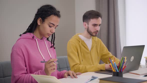 Portrait of Young African American Girl and Caucasian Boy Sitting at the Table and Studying alt
