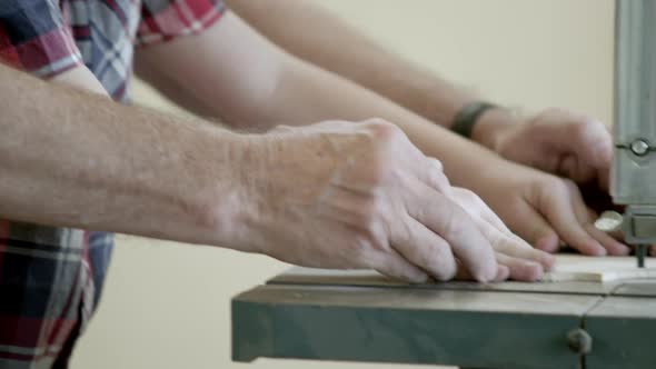Tight panning shot of grandfather's hands helping and guiding grandson cut wood with an band saw. alt