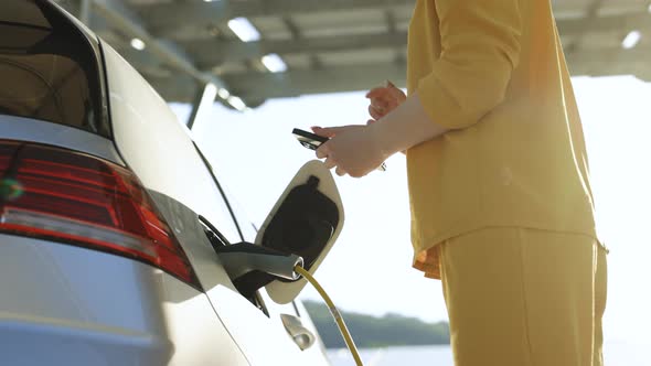 Woman Plugging In an Electric Car at Outlet Socket Activates Charging App on Phone
