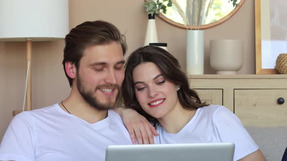 Happy young couple sitting on couch at living room and looking at camera during video chat. alt