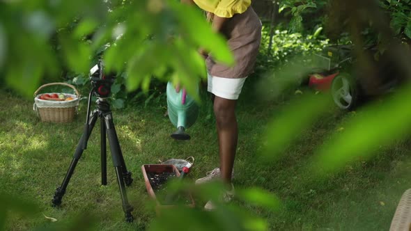 Unrecognizable African American Blogger Watering Seedlings in Pot Filming Video Blog in Garden alt