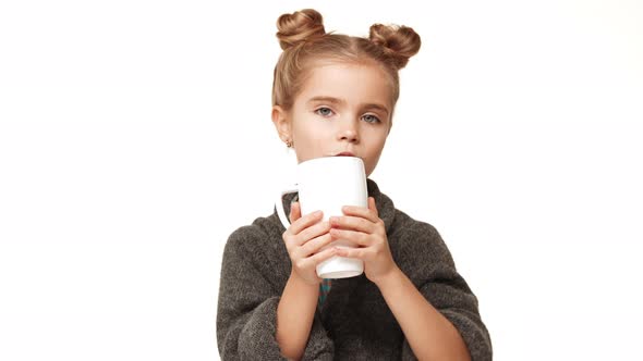 Small Charming Caucasian Kid Girl with Hair Buns and Cozy Woollen Sweater Drinking From Cup and alt