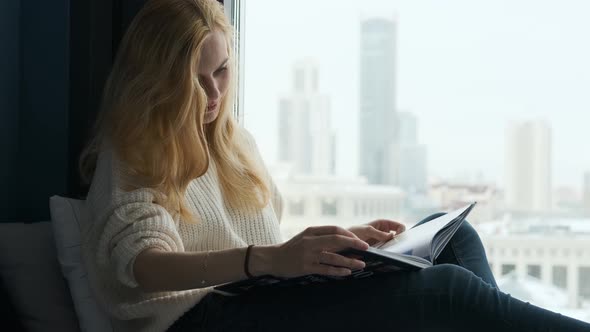 woman reading a book sitting by a large window