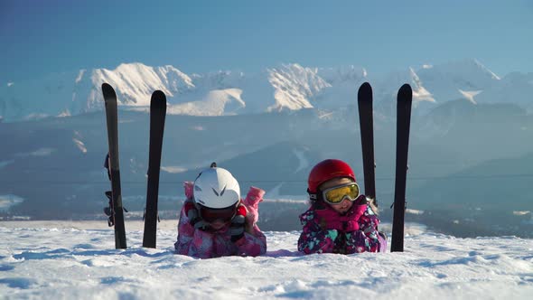 Children in Helmets and Goggles Lying on Snowy Terrain with Skis Near on Background of Mountains