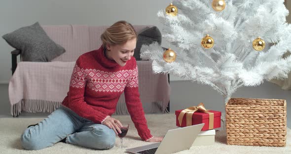 Caucasian Young Woman in Red Sweater Sits at Home on Floor Near Christmas Tree Drinks Wine From alt