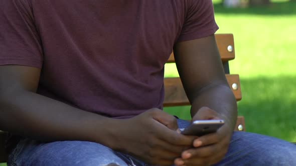 Concentrated Afro-American Man Sitting on Bench in Park, Using His Smartphone alt
