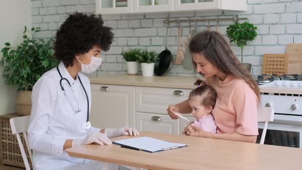 Afro American Woman Doctor in Mask Checks Baby's Body Temperature at Home Next to Mom alt