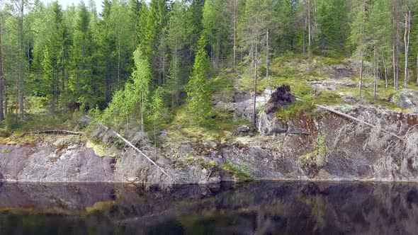 Drone sliding in front of a steep cliff above a calm forest lake in the boreal wilderness. Fallen pi alt
