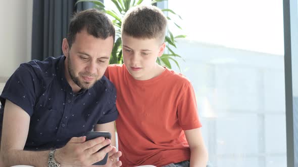 Father and Teenage Son Watching Videos on the Phone While at Home alt