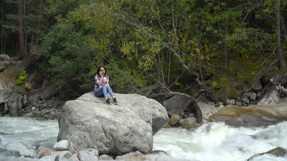 A Young Woman Sits on a Stone and Speaks on the Phone in the Mountains By the River. Travel To the alt