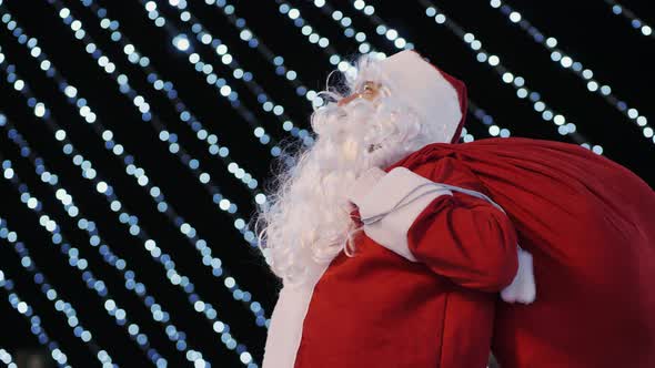 Santa Claus Looks Up at the City Christmas Tree and Bright Flashing Lights of Garlands alt