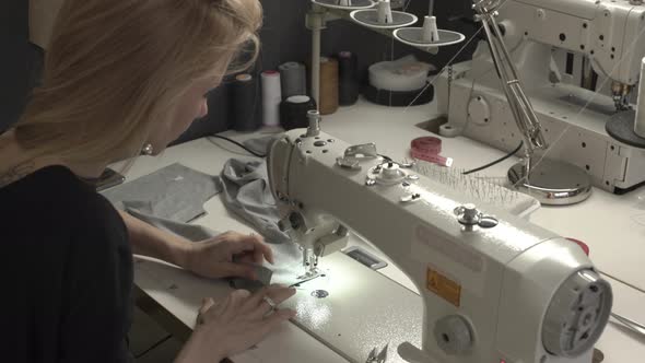 Blonde Caucasian Female in Black Dress Sitting at Table with Machine in Sewing Classroom Stitching alt