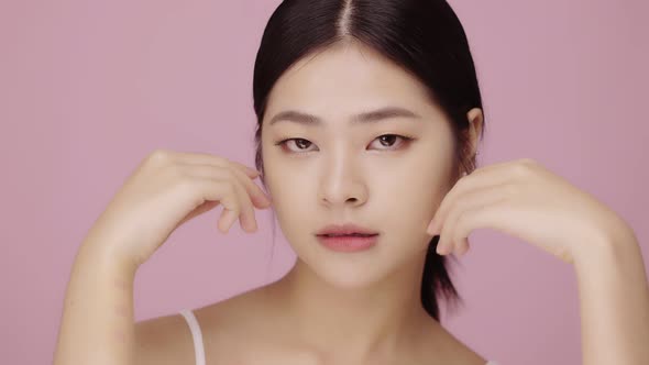 Close up face of beautiful young Asian woman with clear skin lifting face on pink background.
