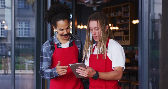 Two diverse male baristas wearing aprons standing in the doorway and using digital tablet alt