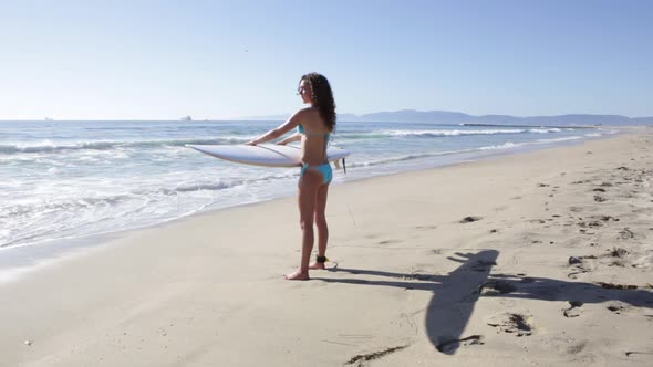 A young woman surfer standing on the beach holding her surfboard as she looks out towards the ocean alt