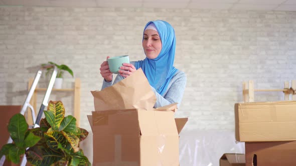 Young Muslim Woman in a Hijab Disassembles a Box of Dishes After Moving alt