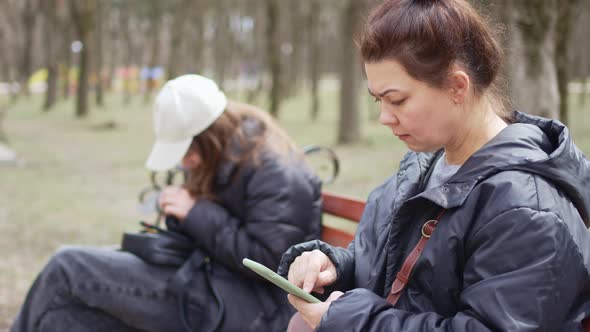 a Woman and a Girl in Black Jackets Sit on a Bench with Smartphones alt