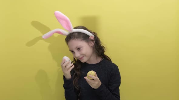 Little Girl in Easter Bunny Ears Holding Two Painted Easter Eggs on a Yellow alt