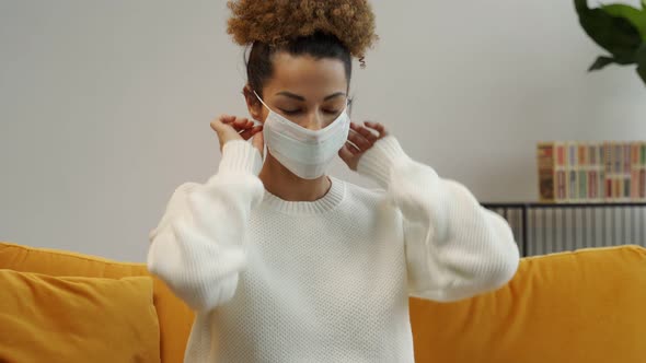 A Black Woman Puts a Mask on Her Face While Sitting on a Yellow Sofa at Home During a Virus Pandemic alt