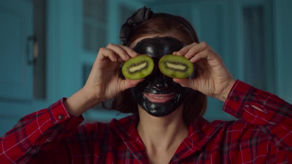 30s woman with black cosmetic mask on her face holding kiwi fruit halves near eyes alt