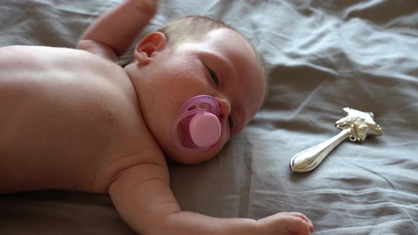 Crying Newborn Baby Child on a Bed with an Iron Silver Rattle and a Pacifier alt