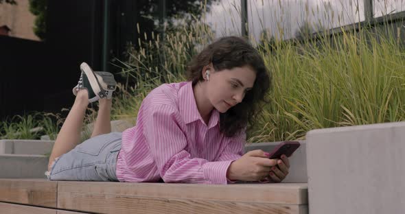 Smiling white woman in city, lying on deck wearing headphones and using smartphone in park. alt