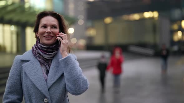 a Woman in a Light Coat and Scarf and with Bright Nails is Talking on the Phone and Laughing at Dusk alt