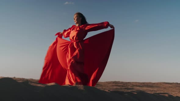 An Asian woman in a red dress whirls on sand dunes alt