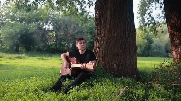 A Young Guy a Guitarist Plays an Acoustic Guitar Near a Tree in a Summer Park Sitting on the Grass