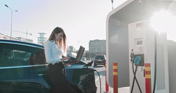 Business Woman Holding Laptop Near Electric Car Car Charging Station alt