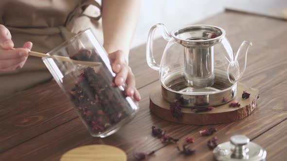 Red Tea Brewing Process. Step One. Housekeeper Sprinkles Dry Hibiscus Tea From Glass Jar With Spoon alt