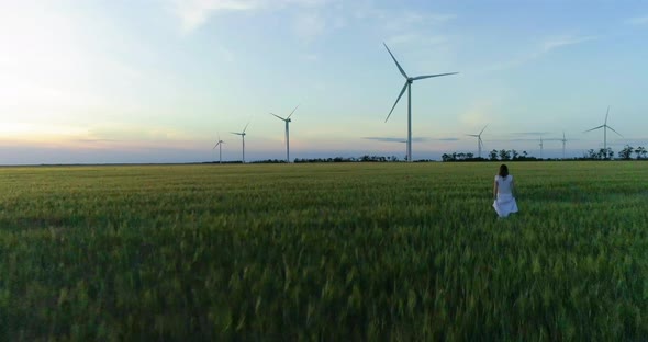 Beautiful girl walking on a green wheat field with windmills for electric power production alt