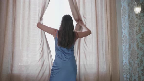 Young Woman Approaches the Window in the Bedroom Pushes Beige Curtains with Her Hands and Enjoys