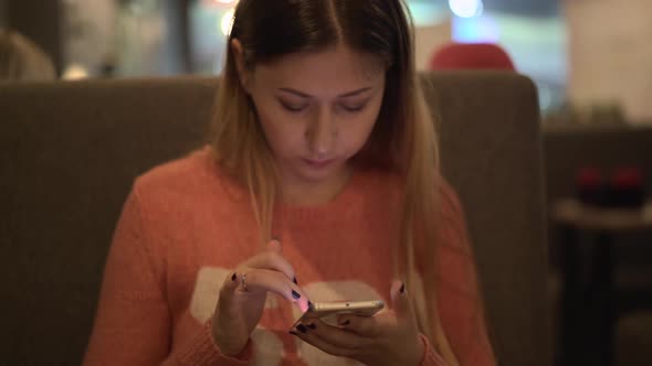 Young Beautiful Girl Sits in a Cafe Works on a Smartphone alt