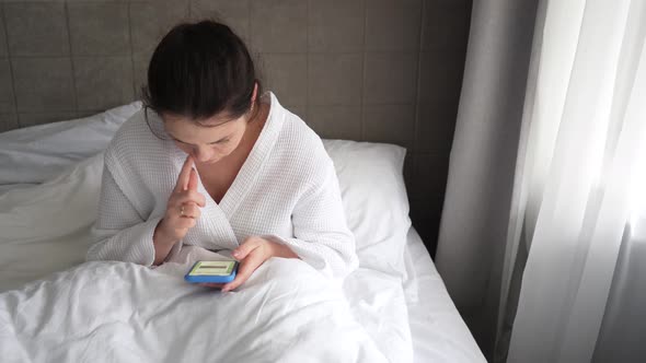 Young Woman in the Morning Lying in Bed and Drinking Coffee Looking at Messages on the Phone