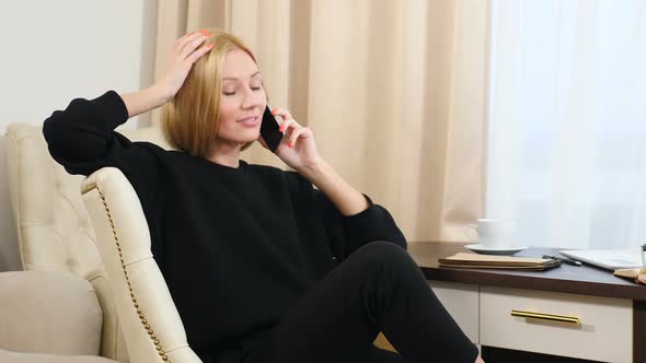 Beautiful Young Woman in Black Sits in Luxury Hotel Room and Talks on Phone alt