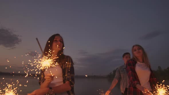 Cheerful Male and Female Friends are Running Along the Beach at Sunset Holding Sparkling Fireworks alt