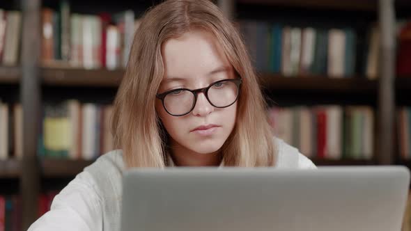Studying Online Teen Girl Wearing Glasses Looking Laptop Typing Message Over Library Background alt
