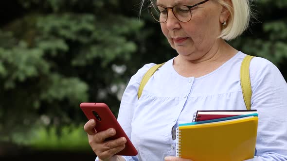 Adult Female Teacher in Eyeglasses with Books and Notebooks Uses a Smartphone While Standing on the