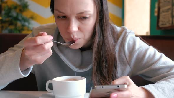 Woman Is Drinking a Hot Chocolate with Spoon and Look on the Screen of Mobile Phone