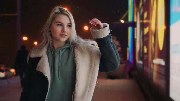 Girl Walks and Smiles Winter Night During Snowfall Near Glowing Shopping Center alt