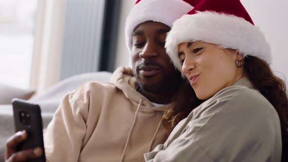 Young Smiling Couple Wearing Santa Hats At Home Posing For Christmas Selfie On Mobile Phone Together