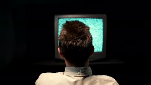 Rear View of a Man Sitting in Front of an Old TV with Noise Interference on a TV Monitor in a Dark alt
