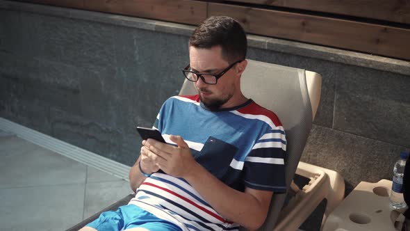 Adult Man Is Resting on Lounger in Yard of Cottage, Using Smartphone alt