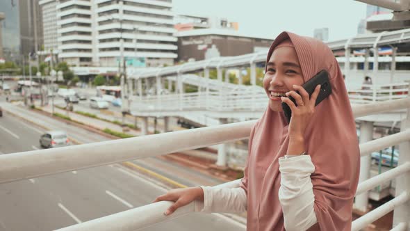 A Young Muslim Girl in a Pink Hijab Talking on the Phone Above Road Traffic in the City Center alt