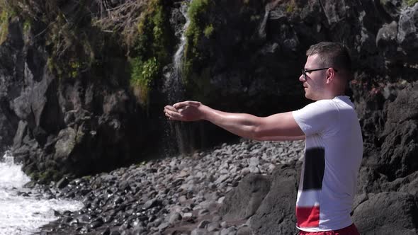 A man on the beach pouring the black sand from his hands under a waterfall alt