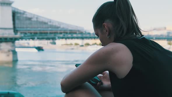 Young Woman Uses Smartphone Outdoor on the Embankment in the Park Near River alt
