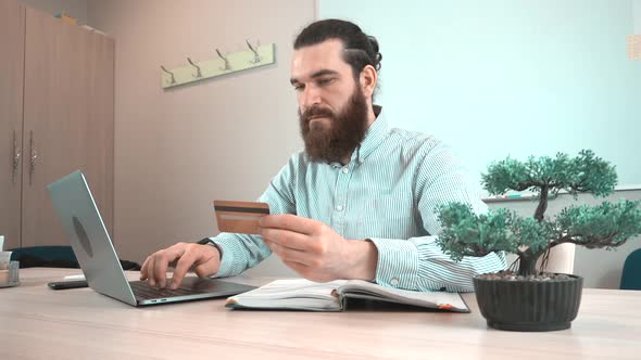 Footage of young bearded man in office making a payment by credit card at laptop alt