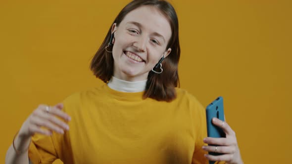 Slow Motion Portrait of Happy Lady Wearing Earpods and Holding Smartphone Dancing on Yellow alt